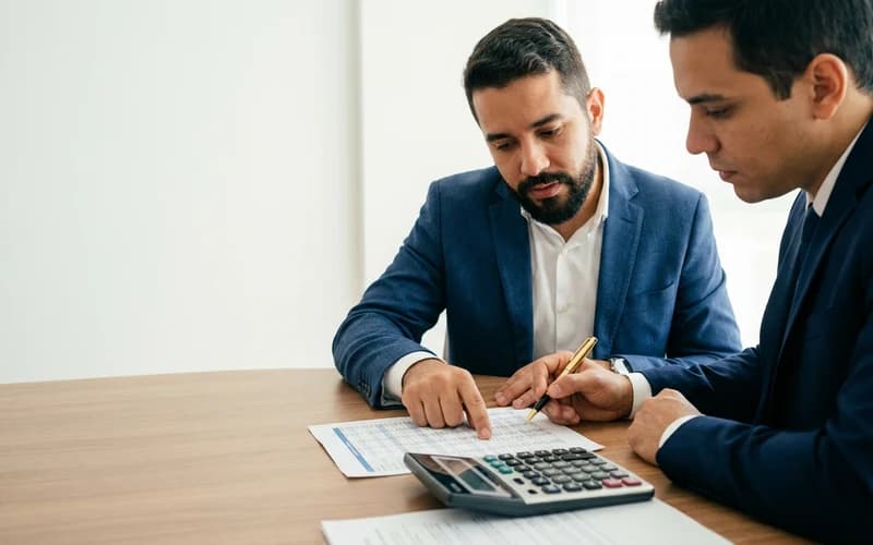 Dois empresários conferem uma planilha de números com calculadora sobre a mesa